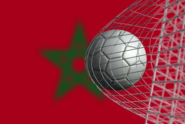 A silver soccer ball hitting the back of a goal net with a blurred Moroccan flag featuring a green star on a red background in the distance.