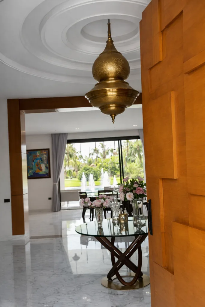 Interior view of an off-market luxury villa Marrakech featuring a grand brass chandelier, marble floors, and a view of the private garden with a pool.